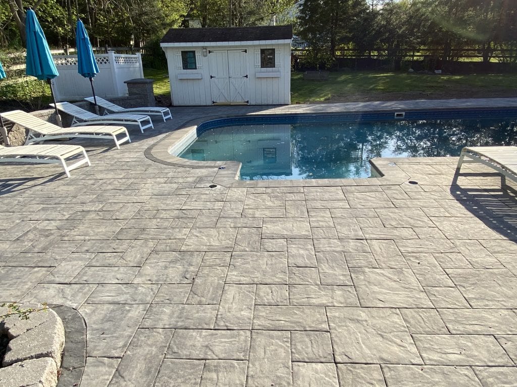A backyard pool area features a gray stone patio, expertly crafted by a hardscaping contractor. Several white lounge chairs and blue umbrellas add comfort, while a small white shed nestles in the background, surrounded by trees and lush green lawn.
