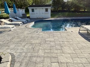 A backyard pool area features a gray stone patio, expertly crafted by a hardscaping contractor. Several white lounge chairs and blue umbrellas add comfort, while a small white shed nestles in the background, surrounded by trees and lush green lawn.