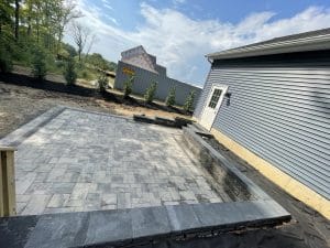 A newly constructed stone patio next to a gray house, bordered by a low stone wall, showcases the craftsmanship of a skilled hardscaping contractor. The surrounding area boasts fresh soil and trees in the background, with a partly cloudy sky visible above.
