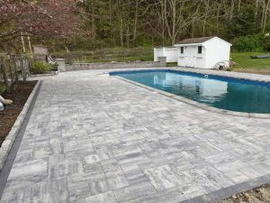 The backyard scene features a rectangular swimming pool surrounded by expertly installed gray stone pavers, courtesy of professional patio installation. A small white shed stands in the background, with trees creating a natural border. The open area boasts a neat and clean layout, enhancing the serene ambiance.