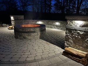 Concrete paver patio with fire pit and outdoor lighting shown at night.