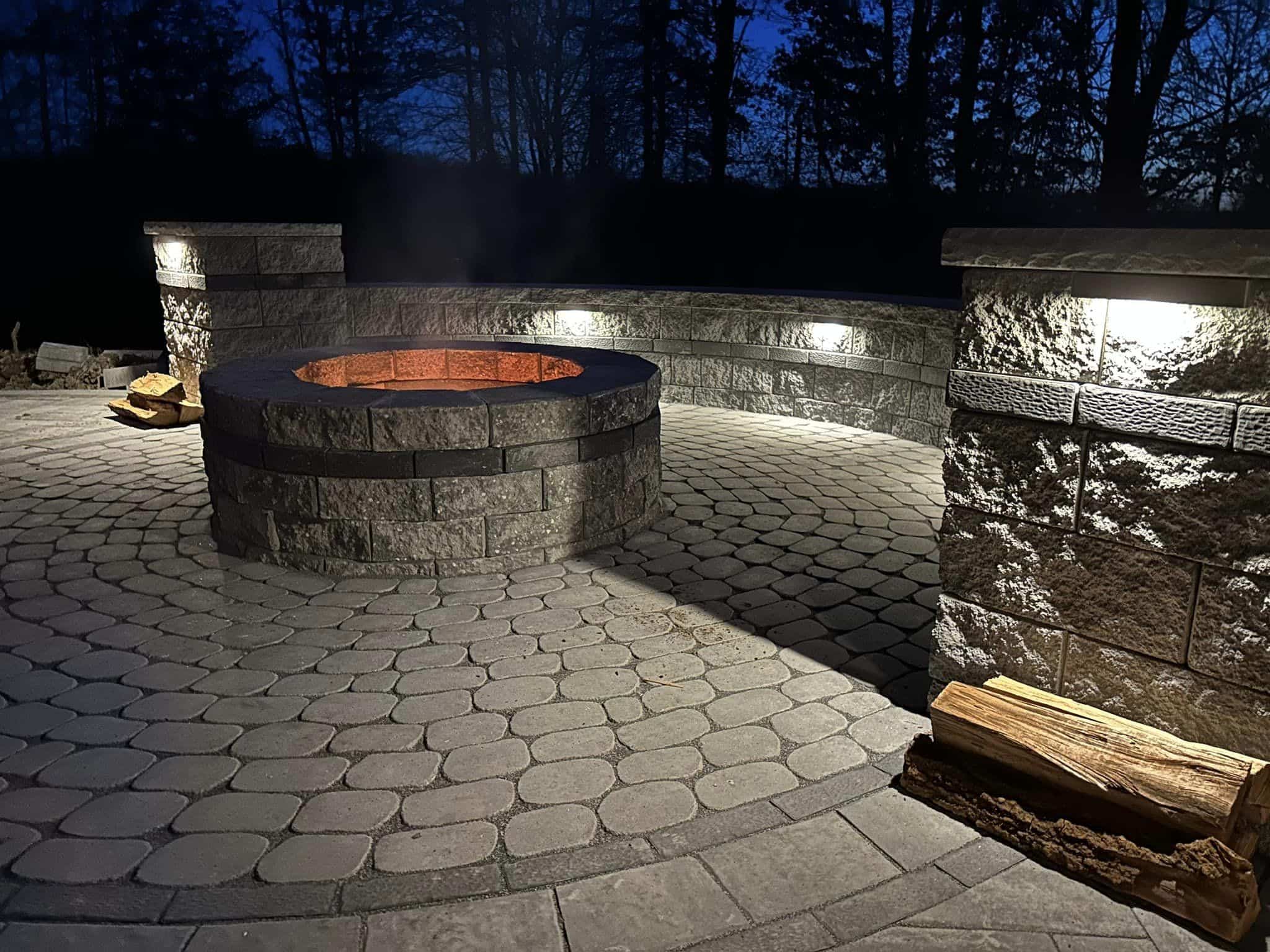 Concrete paver patio with fire pit and outdoor lighting shown at night.