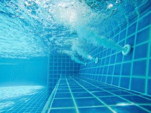 Underwater view of a swimming pool with blue tiles and water jets creating bubbles. Sunlight filters through the water, highlighting the texture of the tiles and the motion of the bubbles.