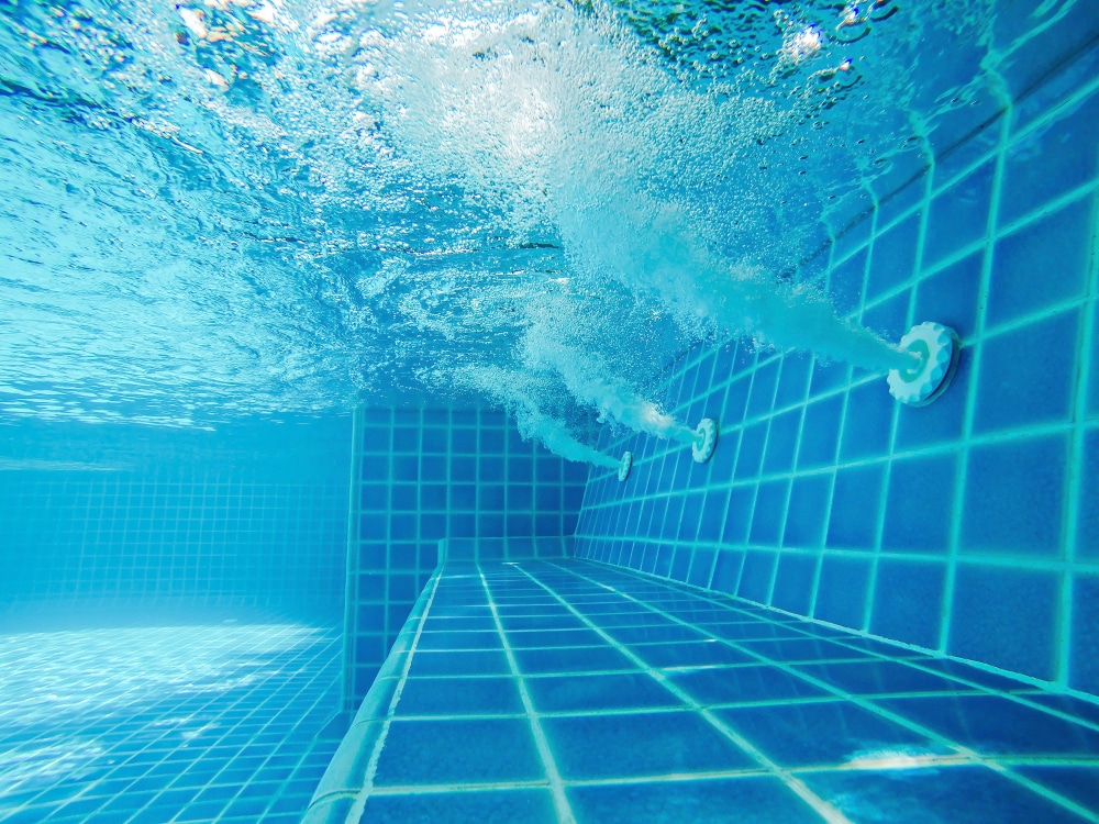 Underwater view of a swimming pool with blue tiles and water jets creating bubbles. Sunlight filters through the water, highlighting the texture of the tiles and the motion of the bubbles.