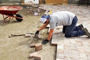 A person in a cap and gloves is kneeling on the ground, placing paving stones to construct a patio. Nearby, a wheelbarrow and a shovel rest on the sandy surface.