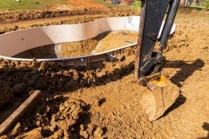 A construction site with a partially installed in-ground swimming pool. An excavator is parked beside the pool, with its arm and bucket resting on the muddy ground. The area is surrounded by dug-up dirt and soil.