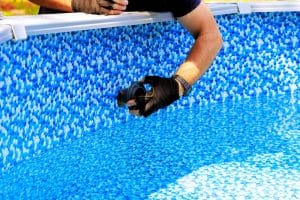A person wearing black gloves is installing a jet fitting into a blue above-ground pool. The interior of the pool features a blue and white mosaic pattern. The person is leaning over the pool's edge, focusing on their task.