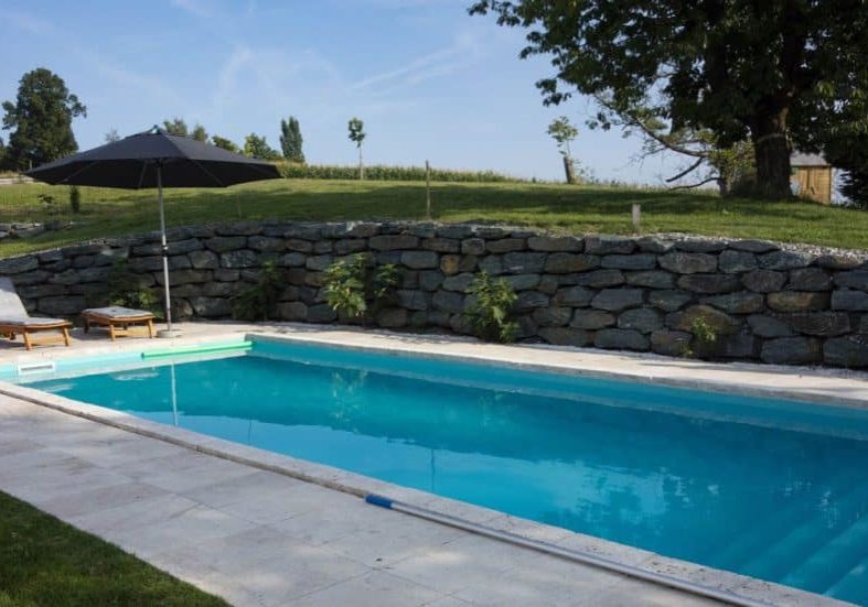 Fiberglass pool with stone deck and large natural stone retaining wall in background.